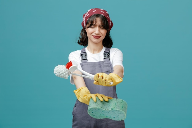 Jeune femme nettoyante mécontente portant un bandana uniforme et des gants en caoutchouc étirant une éponge et des brosses de toilette vers la caméra en regardant des brosses isolées sur fond bleu