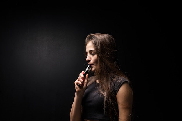 Jeune femme en noir fume une cigarette électronique sur un mur sombre