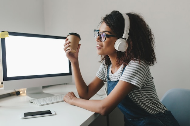 Jeune femme noire en détournant les yeux avec nostalgie tenant une tasse de café et souriant tout en travaillant au bureau