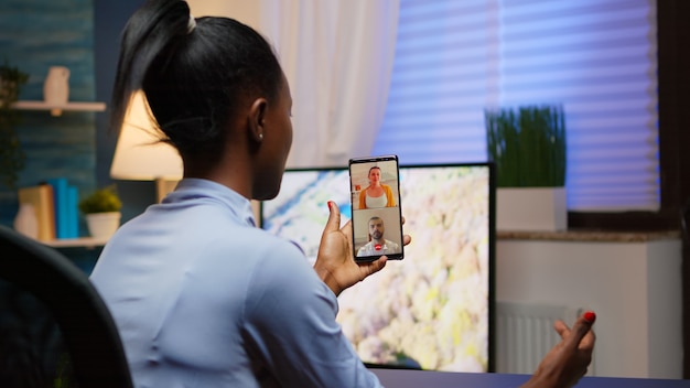 Jeune femme noire discutant sur webcam avec sa famille à l'aide d'un smartphone faisant une pause après avoir fait des heures supplémentaires à la maison. Indépendant travaillant sur un lieu de travail personnel ayant une réunion vidéo.