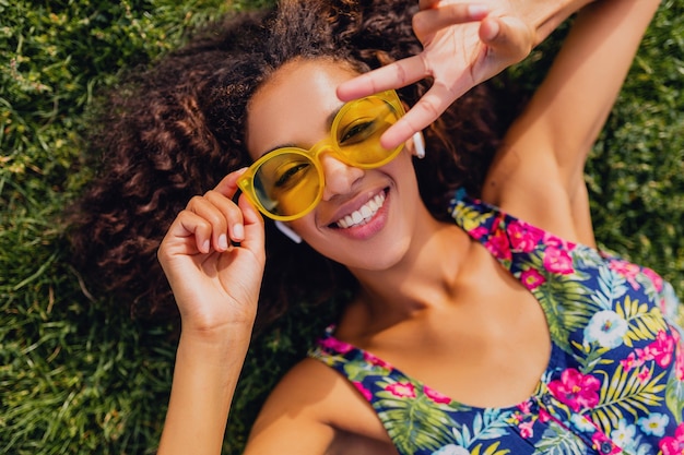 Photo gratuite jeune femme noire élégante, écouter de la musique sur des écouteurs sans fil s'amuser allongé sur l'herbe dans le parc, style de mode d'été, tenue de hipster coloré, vue d'en haut