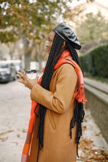 Jeune femme noire avec une longue coiffure à l'extérieur avec une tasse de café à emporter. Femme portant un manteau marron, une écharpe orange et un chapeau noir