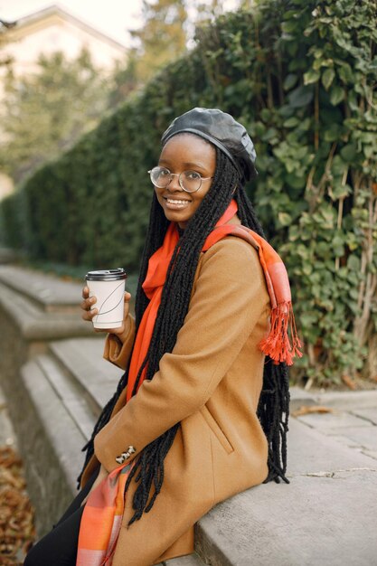 Jeune femme noire avec une longue coiffure locs assis dehors avec une tasse de café à emporter. Femme portant un manteau marron, une écharpe orange et un chapeau noir