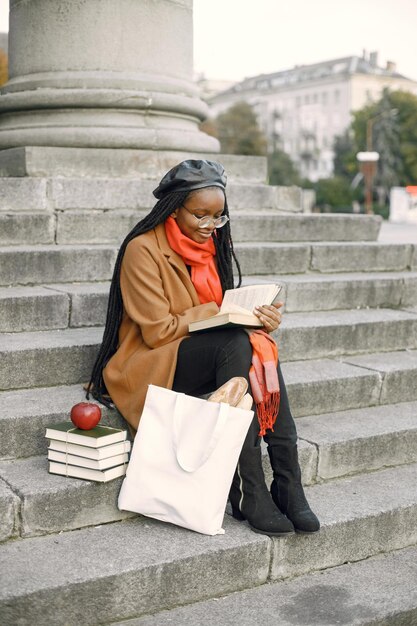 Jeune femme noire avec de longues coiffures locs assise sur un escalier avec un livre. Femme portant un manteau marron, une écharpe orange et un chapeau noir