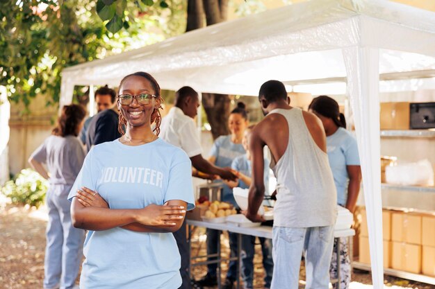 Une jeune femme noire avec des lunettes se tient à l'extérieur, les bras croisés, regardant la caméra. Un groupe diversifié de bénévoles soutient un programme à but non lucratif dédié à la lutte contre la faim et à l'aide aux personnes dans le besoin.