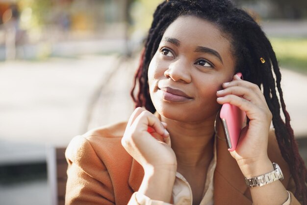 Jeune femme noire positive assise dans un café en plein air et parlant au téléphone