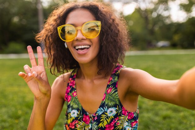 Jeune femme noire positive élégante prenant selfie photo en écoutant de la musique sur des écouteurs sans fil s'amuser dans le parc, style de mode estivale, tenue hipster colorée