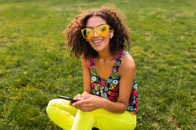Jeune femme noire souriante élégante à l'aide de smartphone, écouter de la musique sur des écouteurs sans fil s'amuser dans le parc
