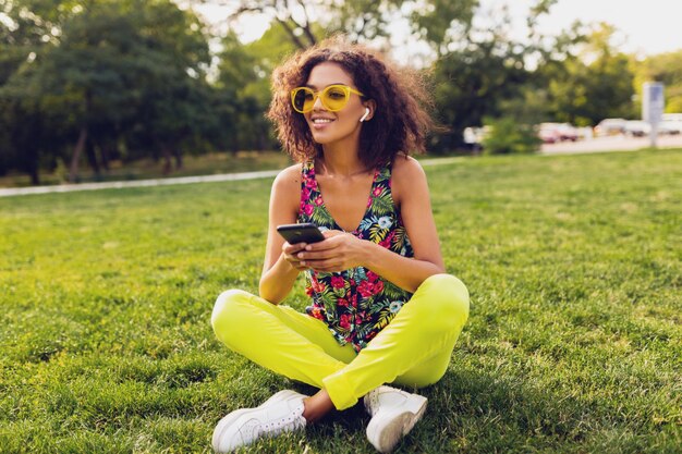 Jeune femme noire souriante élégante à l'aide de smartphone, écouter de la musique sur des écouteurs sans fil s'amuser dans le parc