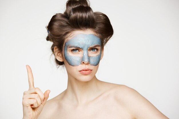 Jeune femme nue en bigoudis et masque facial fronçant les sourcils. Soins de beauté et cosmétologie.