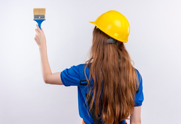 Jeune femme ouvrier constructeur en uniforme de construction et casque de sécurité debout avec son dos tenant un pinceau sur un mur blanc