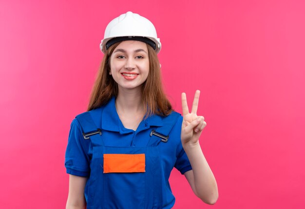 Jeune femme ouvrier constructeur en uniforme de construction et casque de sécurité souriant joyeusement montrant signe de la victoire sur le numéro deux debout sur le mur rose