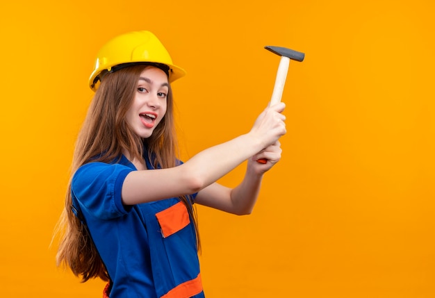 Jeune femme ouvrier constructeur en uniforme de construction et casque de sécurité tenant un marteau souriant joyeusement debout sur un mur orange