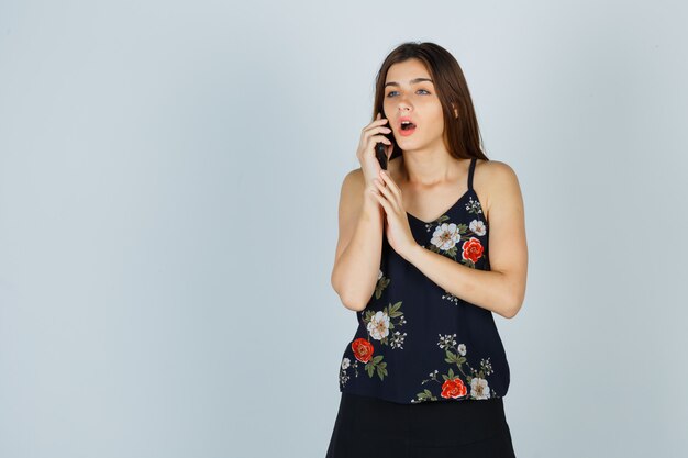 Jeune femme parlant sur smartphone en blouse et à la perplexité, vue de face.