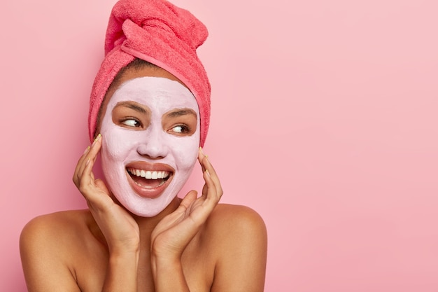 Jeune femme avec une peau foncée fraîche et saine applique un masque d'argile nourrissant, se tient avec les épaules nues, regarde joyeusement loin, a enveloppé une serviette sur la tête, prend un bain, regarde de côté avec une expression joyeuse