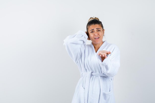 Jeune femme en peignoir blanc étirant la main comme recevant quelque chose tout en tenant la main derrière la tête et l'air ennuyé