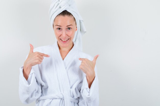 Jeune femme en peignoir blanc, serviette pointant sur elle-même et à la joyeuse vue de face.