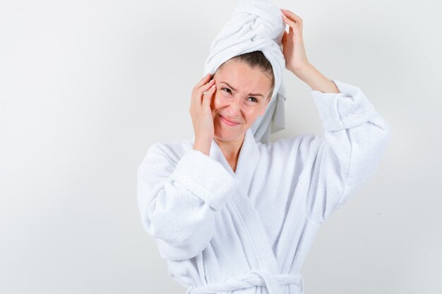 Jeune femme en peignoir blanc, serviette tenant les mains sur la tête et la joue et regardant agité, vue de face.