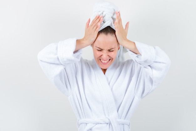 Jeune femme en peignoir blanc, serviette tenant les mains sur la tête et regardant agressif, vue de face.