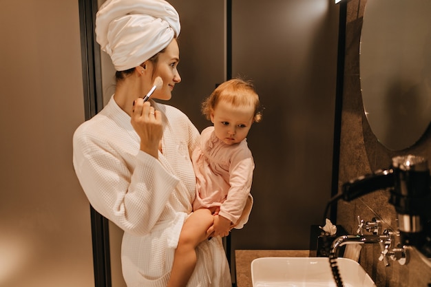 Jeune femme en peignoir détient fille blonde et fait son propre maquillage, à la recherche dans le miroir de la salle de bain.