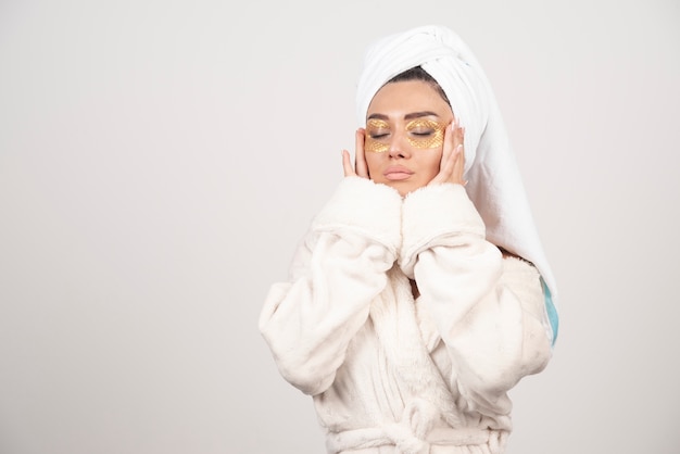 Jeune femme en peignoir et patchs oculaires cosmétiques touchant son visage.