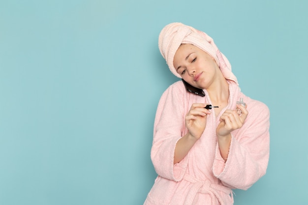 Jeune femme en peignoir rose après la douche parler au téléphone peindre ses ongles sur bleu