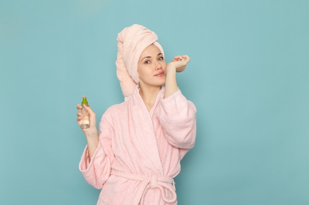 Jeune femme en peignoir rose après la douche tenant et à l'aide de spray sur bleu