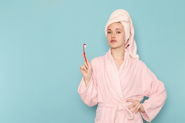 Jeune femme en peignoir rose après la douche tenant la brosse à dents sur bleu