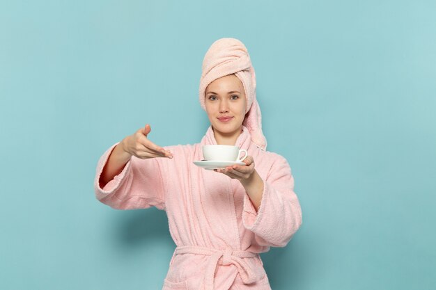 Jeune femme en peignoir rose après la douche tenant une tasse de café avec sourire sur bleu