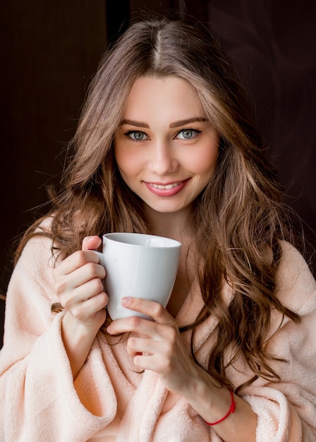 Jeune femme en peignoir tendre rose boire du thé et souriant.