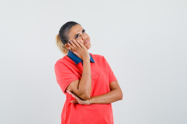 Jeune femme penchée joue sur la paume tout en pensant à quelque chose en t-shirt rouge et à la pensive, vue de face.