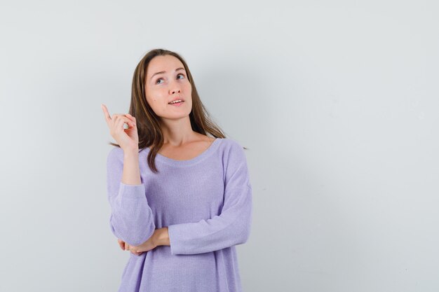 Jeune femme pensant tout en pointant vers le haut en chemisier lilas et à la réflexion. vue de face.