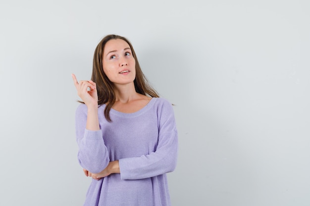 Jeune femme pensant tout en pointant vers le haut en chemisier lilas et à la réflexion. vue de face.