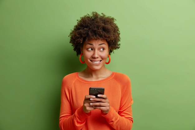 Jeune femme pensive mord les lèvres détient les utilisations cellulaires modernes phoe mobile envoie des messages texte porte cavalier orange et boucles d'oreilles isolés sur mur vert