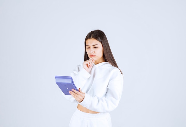 jeune femme pensive regardant une calculatrice sur fond blanc-gris.