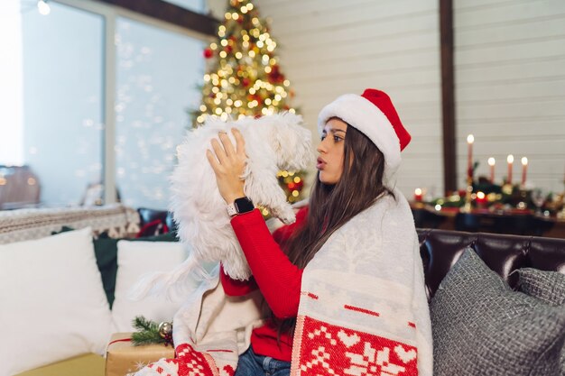 Jeune femme avec un petit chien dans ses bras est assise sur le canapé le soir du Nouvel An