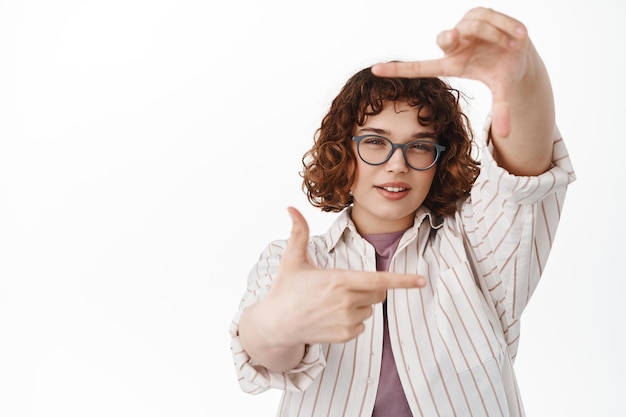 Jeune femme photographe réfléchie et créative créant une image dans son esprit, recherchant un angle parfait pour la photo, regardant à travers le geste des cadres de la main dans les verres, fond blanc