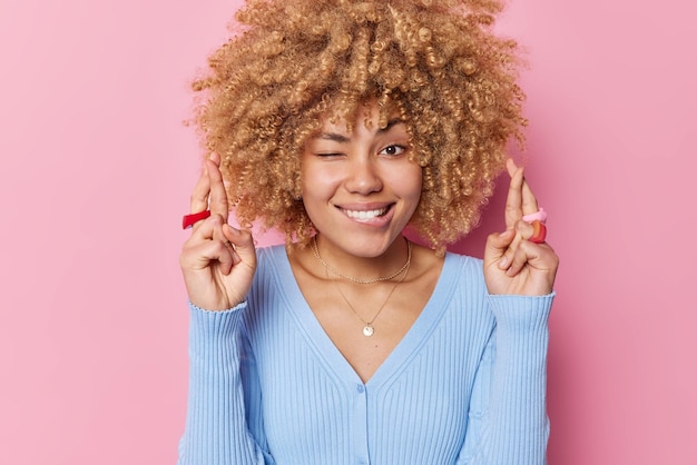 Photo gratuite une jeune femme pleine d'espoir aux cheveux bouclés fait un clin d'œil attend l'annonce des résultats mord les doigts croit en la bonne chance porte un pull bleu décontracté isolé sur fond rose concept de langage corporel