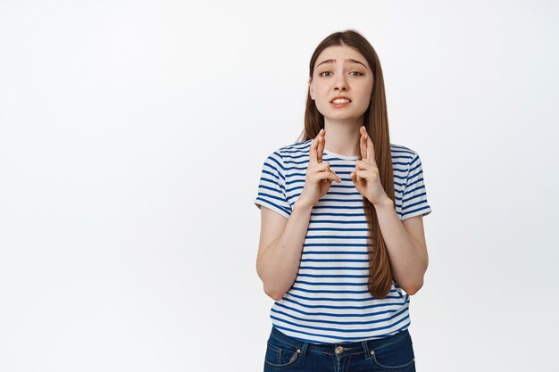 Jeune femme pleine d'espoir debout avec les doigts croisés, espérant que le vœu se réalise, attendant des nouvelles, debout sur blanc.