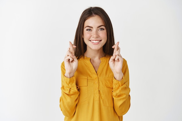 Jeune femme pleine d'espoir faisant un vœu, debout avec les doigts croisés pour la bonne chance, portant un chemisier jaune