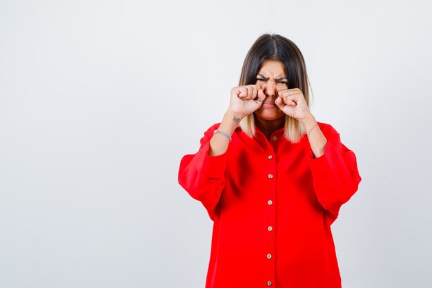 Jeune femme pleurant en se frottant les yeux avec les mains en chemise rouge surdimensionnée et l'air triste, vue de face.