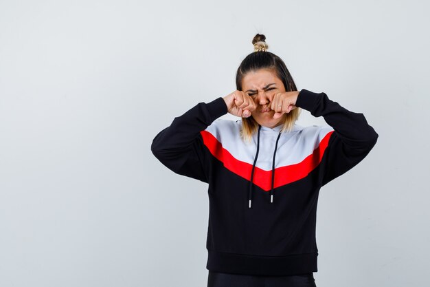 Jeune femme pleurant en se frottant les yeux avec les poings dans un pull à capuche et l'air triste.