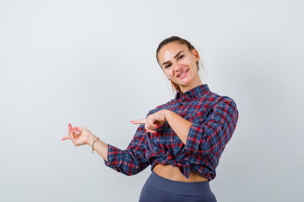 Jeune femme pointant sur le côté en chemise à carreaux et l'air heureux, vue de face.