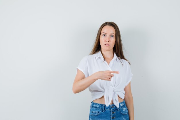 Jeune femme pointant de côté en chemisier blanc et l'air mécontent. vue de face.