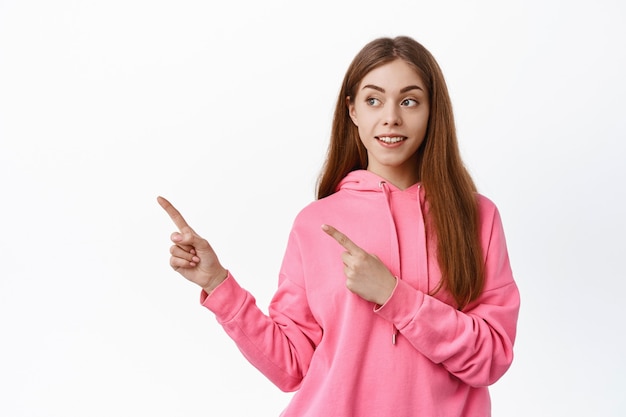 Jeune femme pointant de côté, regardant l'espace de copie gauche avec une expression curieuse, vérifiant la promo, debout contre le mur blanc