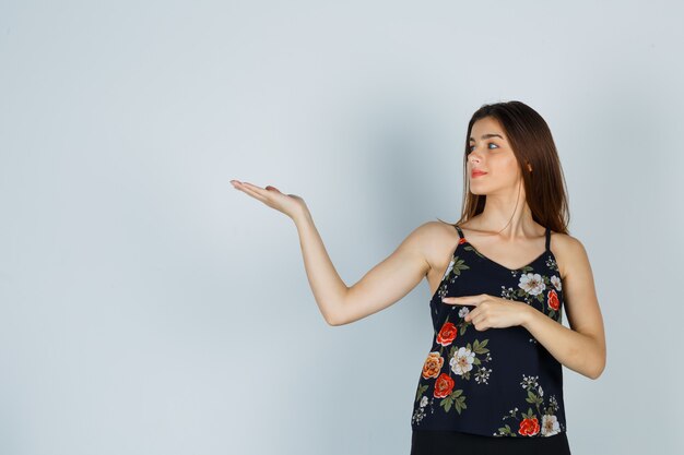 Jeune femme pointant de côté tout en faisant un geste de bienvenue en blouse et en ayant l'air confiant. vue de face.