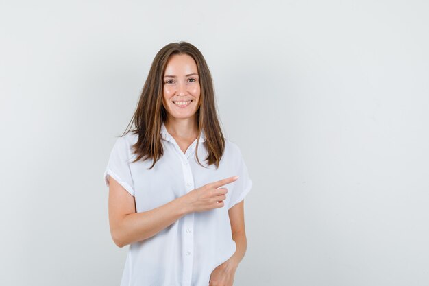 Jeune femme pointant de côté tout en souriant en chemisier blanc et à la confiance