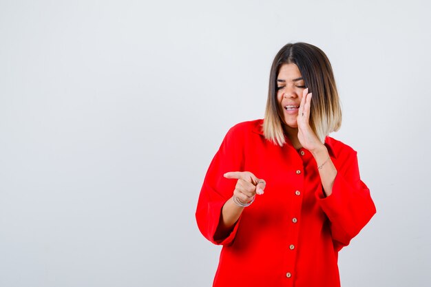 Jeune femme pointant de côté tout en tenant la main sur le côté de la bouche en chemise rouge surdimensionnée et l'air heureux. vue de face.