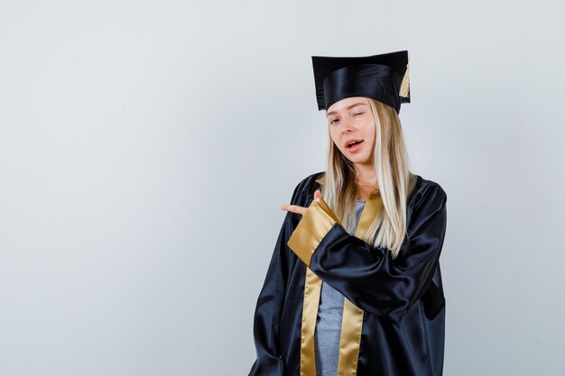 Jeune femme pointant de côté en uniforme de diplômé et ayant l'air confiant.