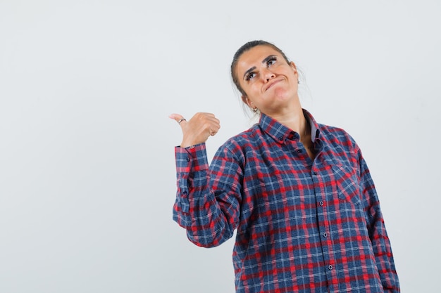 Jeune femme pointant derrière avec l'index en chemise à carreaux et à la jolie. vue de face.